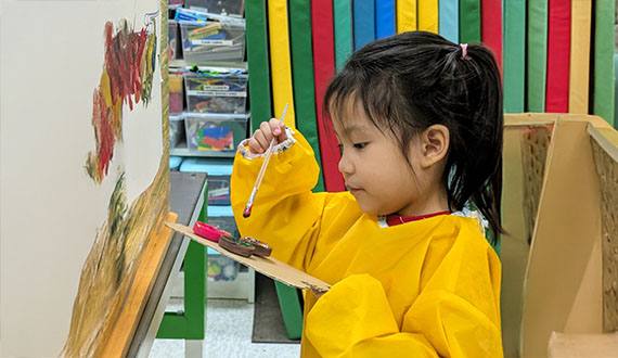 Child in a yellow smock painting at an easel